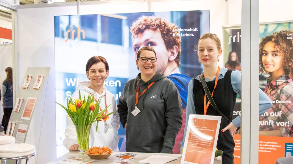 Drei Frauen stehen an einem Messestand, hinter einem Tisch mit Blumen und Informationsmaterialien.