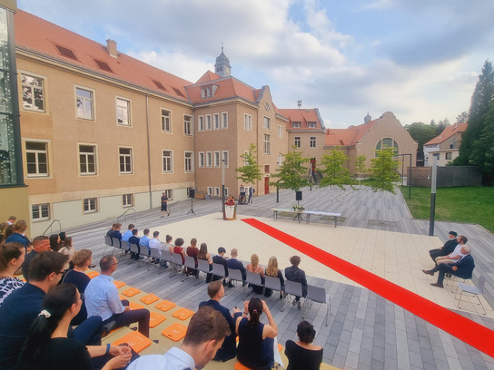Menschen sitzen im Freien vor einem Gebäude, hören einem Redner zu.