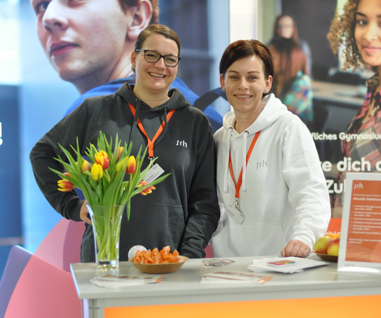 Zwei Personen stehen vor einem orangefarbenen Banner, eine hält Blumen.