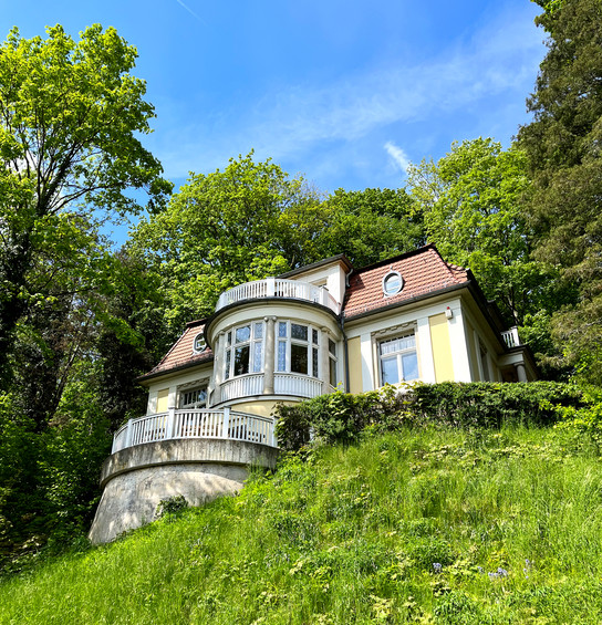 Ein zweistöckiges Haus mit Balkon auf einem grasbewachsenen Hügel, umgeben von Bäumen unter einem klaren blauen Himmel.