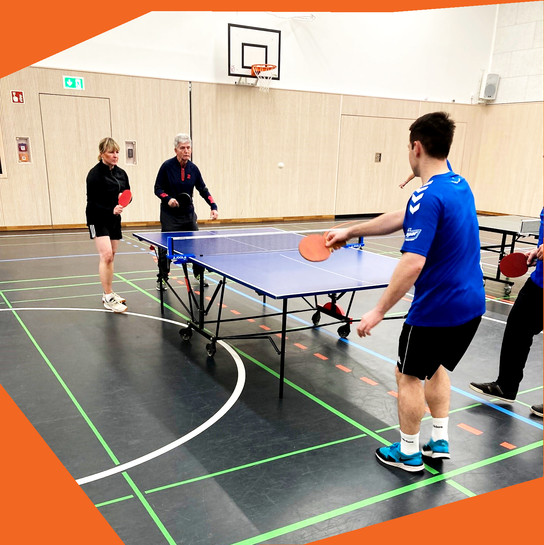 Vier Personen spielen Tischtennis in einem Sporthalleninneren.